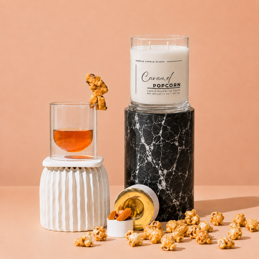 A candle in a clear glass jar labeled 'Caramel Popcorn' by Fragrance Candle Studio, displayed on a black marble pedestal. The scene is styled with caramel popcorn scattered on a peach background, a glass of amber liquid on a white ribbed base, and an open tin with a gold lid.