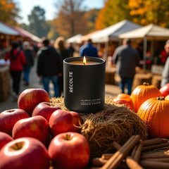 Black candle labeled 'Récolte' surrounded by apples, pumpkins, and cinnamon sticks at an outdoor market.