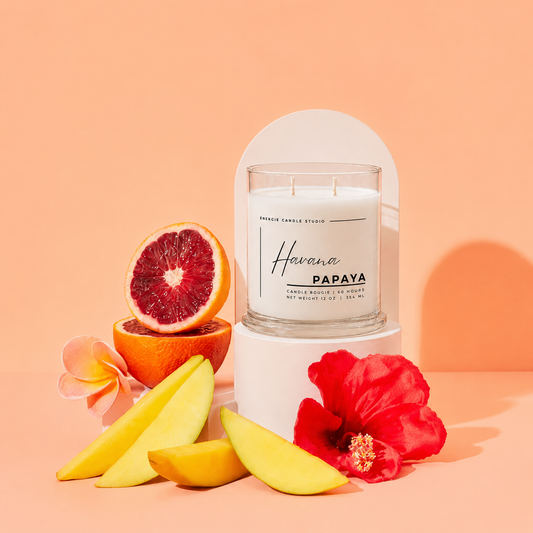 A glass jar candle labeled 'Havana PAPAYA' by Freshly Picked Candle Studio sits on a white pedestal against a peach background, styled with sliced yellow mango, a halved blood orange, a red hibiscus flower, and a pink plumeria.