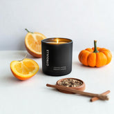 Peppered Pumpkin candle in a black vessel on a white table surrounded by a pumpkin, oranges and a wooden dish of pepper