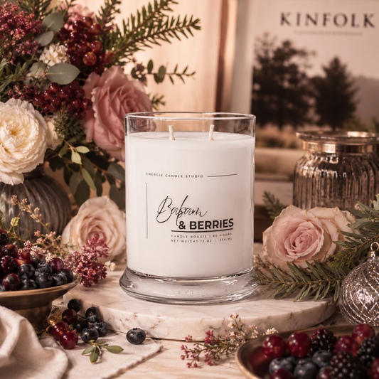 A white scented candle in a clear glass jar sits on a marble slab surrounded by pink roses, blueberries, greenery, and decorative jars. The label reads 'Scented Candle Studio' and 'Blossom & Berries'.