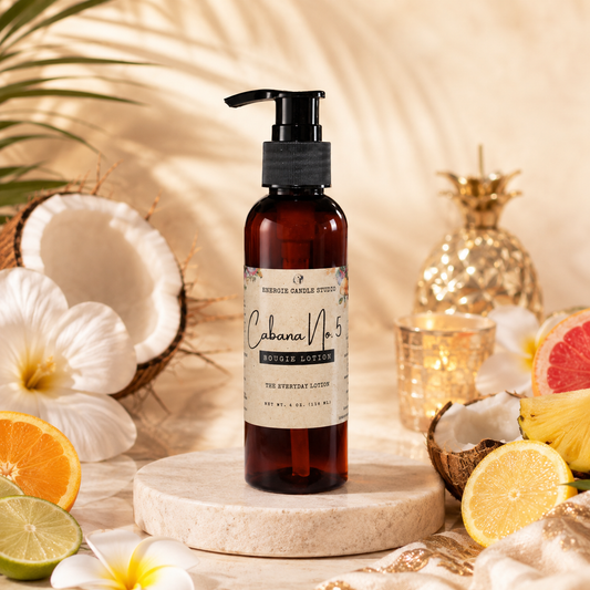 A bottle of Cabana No. 5 Coconut Aromatic room spray stands on a round stone pedestal, surrounded by tropical props including coconuts, flowers, citrus slices, and a gold pineapple against a warm beige background.