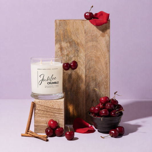 A lit scented candle in a clear glass jar sits on a wooden block, styled with fresh cherries, cinnamon sticks, and red rose petals around a tall wooden pedestal against a lavender background. The candle label reads 'Jubilee CRUMBLE' and 'Tennessee Cherry Wood'.
