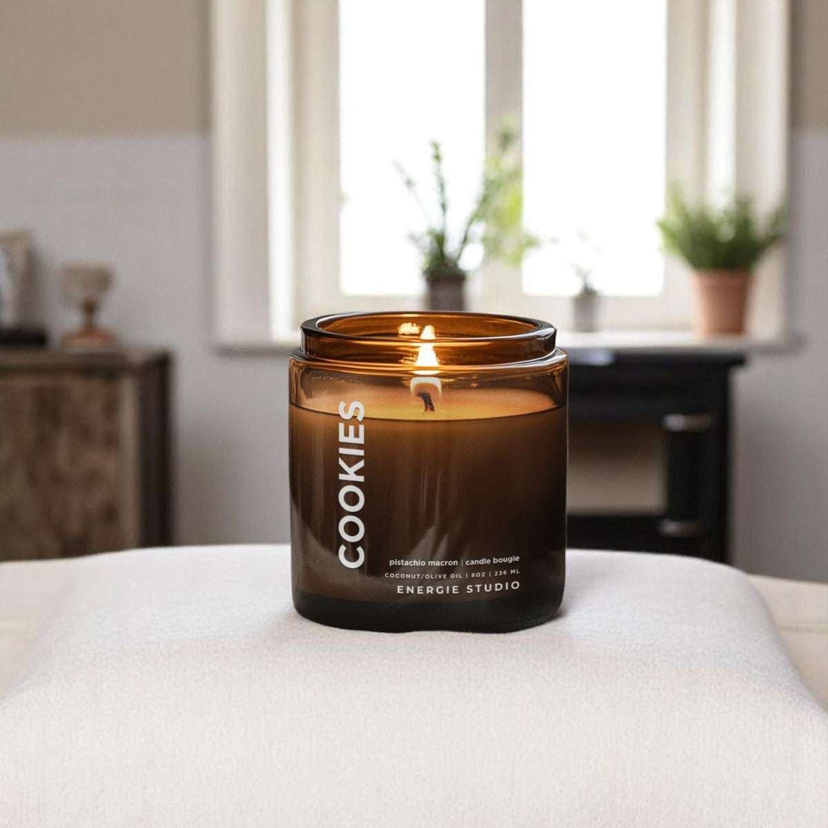 a n amber colored candle sitting on a white blanket with a bedroom behind it with a window and green plants in the background