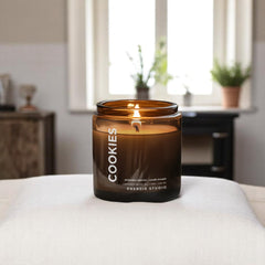 a n amber colored candle sitting on a white blanket with a bedroom behind it with a window and green plants in the background