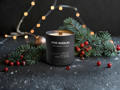 Candle with 'Pine Needles' label surrounded by Christmas decorations on a dark surface.