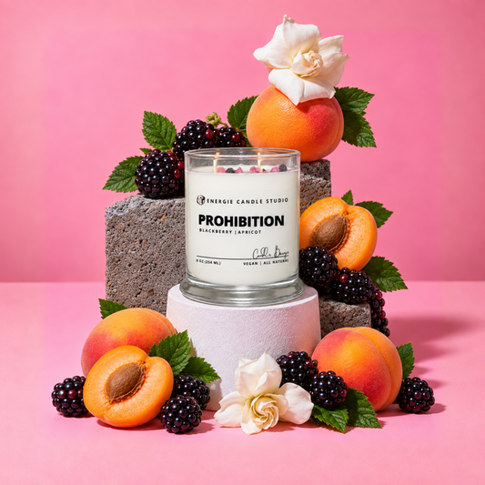 A glass jar candle labeled 'ENERGY CANDLE STUDIO PROHIBITION BLACKBERRY/APRICOT' is styled on a pink background with apricots, blackberries, mint leaves, and white flowers around it.