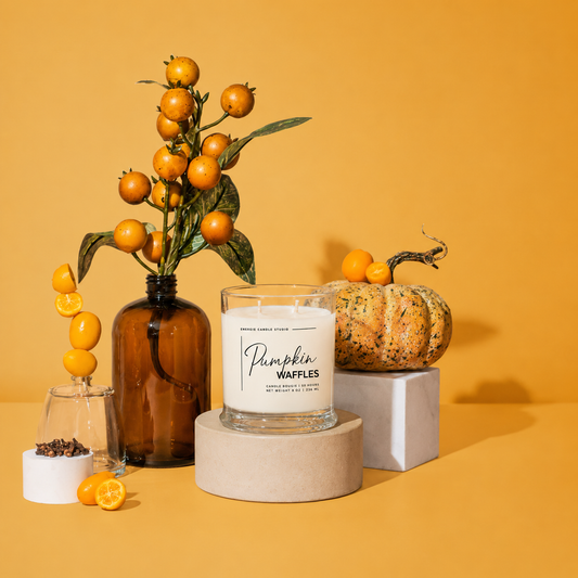 A styled product photo of a candle in a clear glass jar with a white label reading 'Pumpkin Waffles' and 'Peachy Lemon Meringue'. The candle is displayed on a round cardboard pedestal, surrounded by autumn props including an amber glass bottle with a citrus branch, a small pumpkin, and orange slices, all set against a solid mustard yellow background.