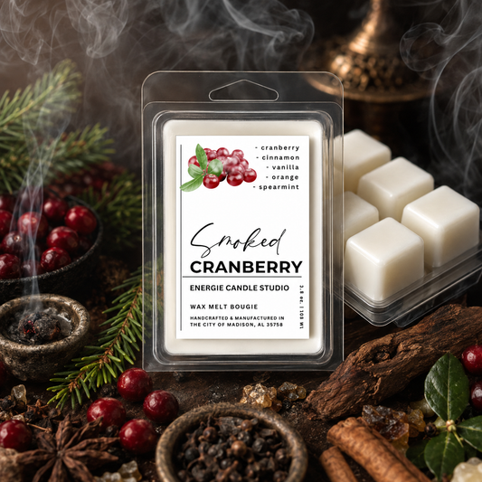 A packaged wax melt candle labeled 'Smoked Cranberry' by Energie Candle Studio, shown in a clear clamshell package with white wax cubes visible. The product is styled with cranberries, evergreen branches, a smoky mug, cinnamon sticks, and dark wood on a rustic surface.