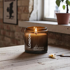 a candle with tobacco and vanilla in London sitting on a wood shelf with an open window and a plant in the background-
