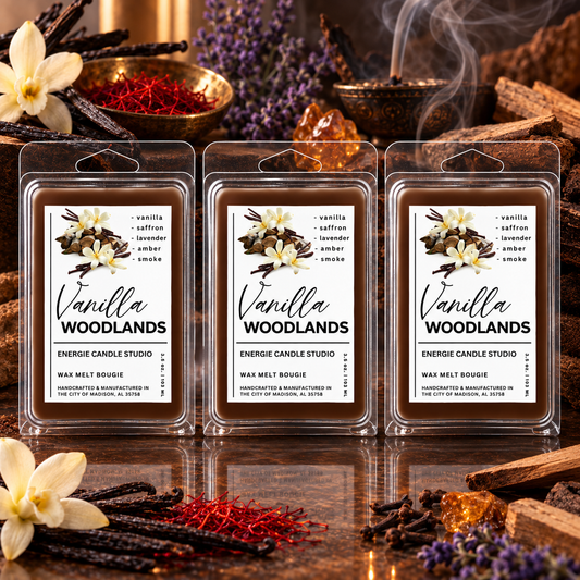 Three packaged wax melts labeled Vanilla Woodlands are arranged on a reflective surface with warm decorative props including flowers, cinnamon sticks, a bowl of red spice, and a copper mug with steam. The product packaging is clear plastic clamshell with a white label featuring vanilla and saffron illustrations.