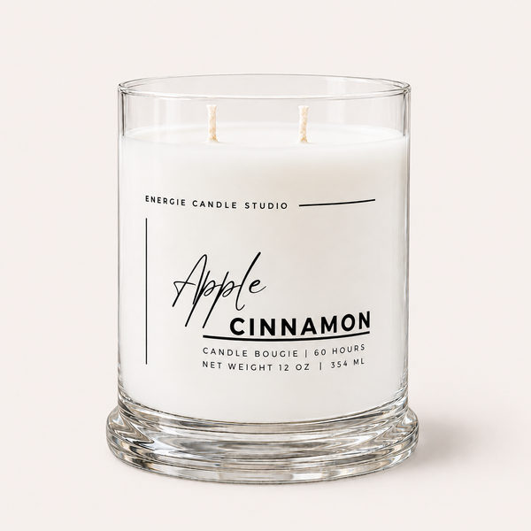 A two-wick scented candle in a clear glass jar with white wax, labeled 'Apple Cinnamon' by Energie Candle Studio, on a light beige background.