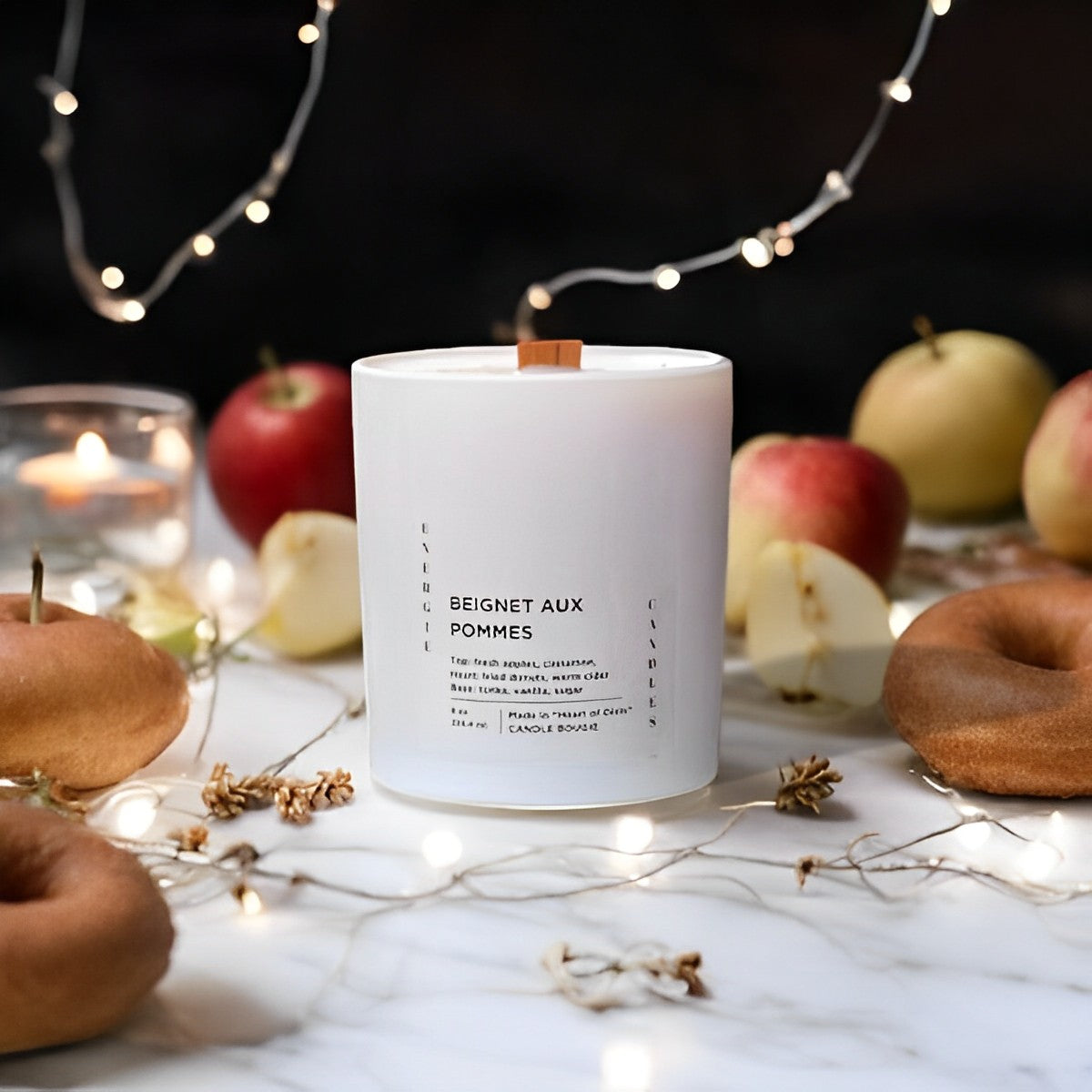 White candle with a wooden wick, placed on a marble table and surrounded by juicy red apples and brown frosted donuts