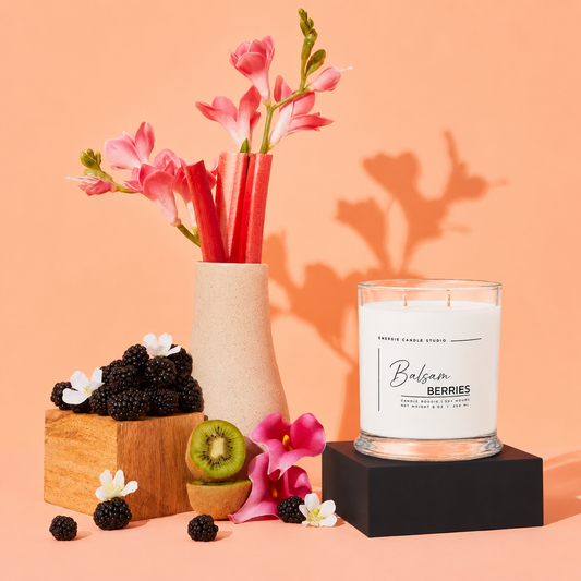 A scented candle in a clear glass jar sits on a black pedestal against a peach studio background. The candle label reads 'Balsam Berries' and 'FRESH BERRY CANDLE STU.