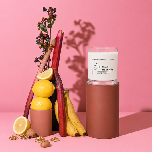 A glass jar candle with white wax sits on a brown cylindrical sleeve against a pink studio background. The candle is styled with lemons, bananas, rhubarb, and walnuts, and the label reads 'Bourbon Nut Bread' with 'Bourbon Candle Co.