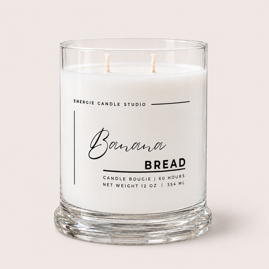 A two-wick candle in a clear glass jar with a thick base, featuring a white wax label that reads 'Banana BREAD' and 'ENERGIE CANDLE STUDIO' in black text, set against a solid light beige background.