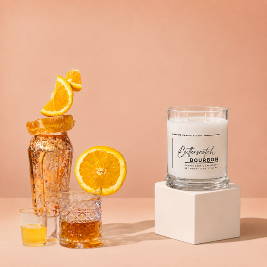 A scented candle in a clear glass jar sits on a white pedestal against a warm peach background. The candle label reads 'Buttercatch Bourbon.