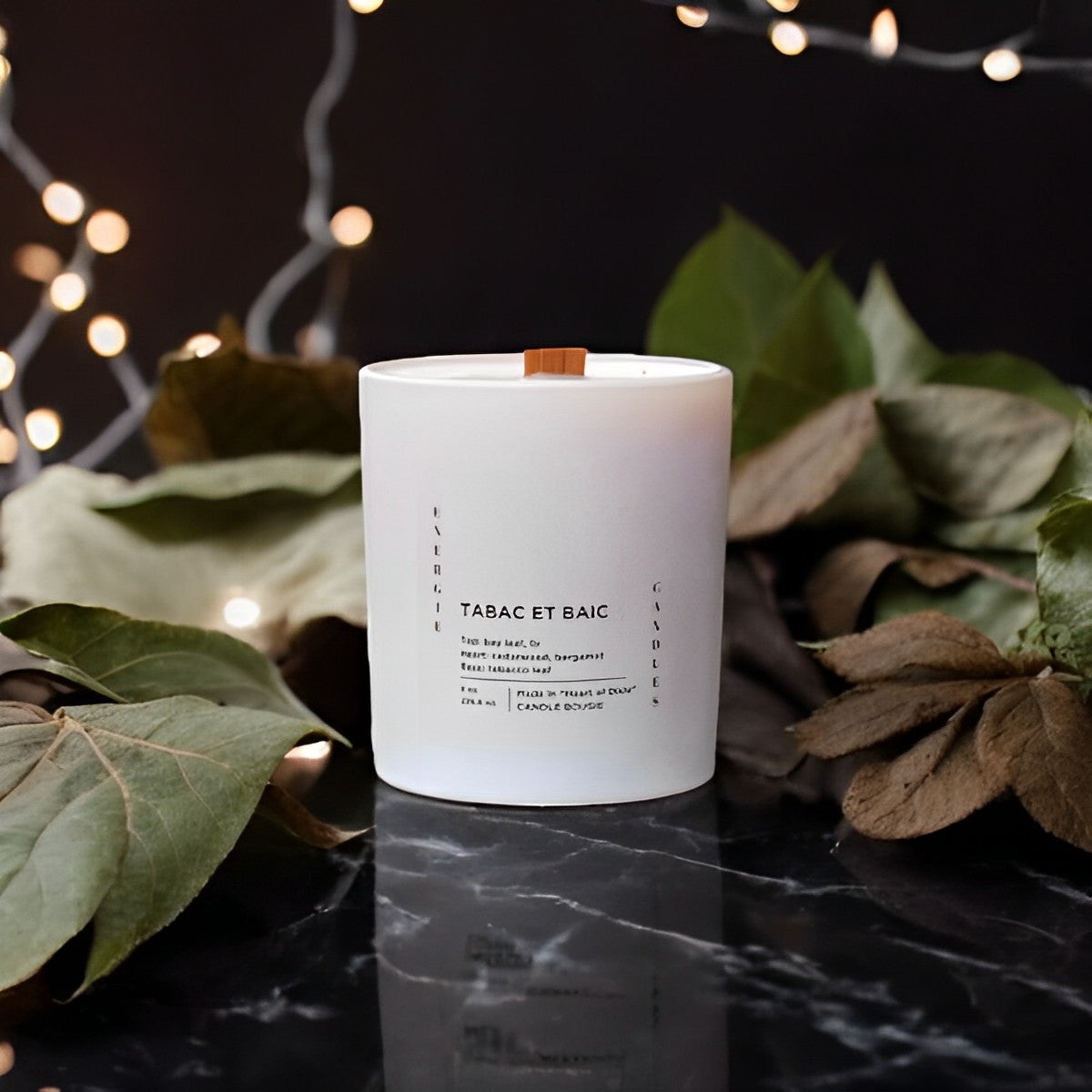 White candle with a wood wick, placed on a black marble table surrounded by bay leaves and string lights, with a black wall in the background.