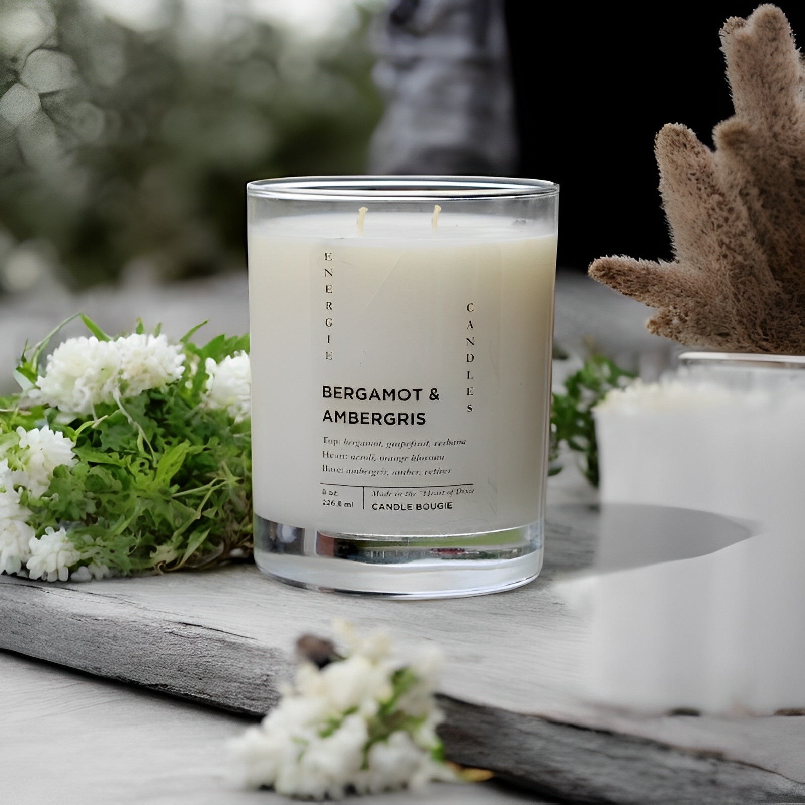 White candle in a clear glass vessel, sitting on a brown wood slate surrounded by white flowers and dried brown flowers  with a gray  blurred background.