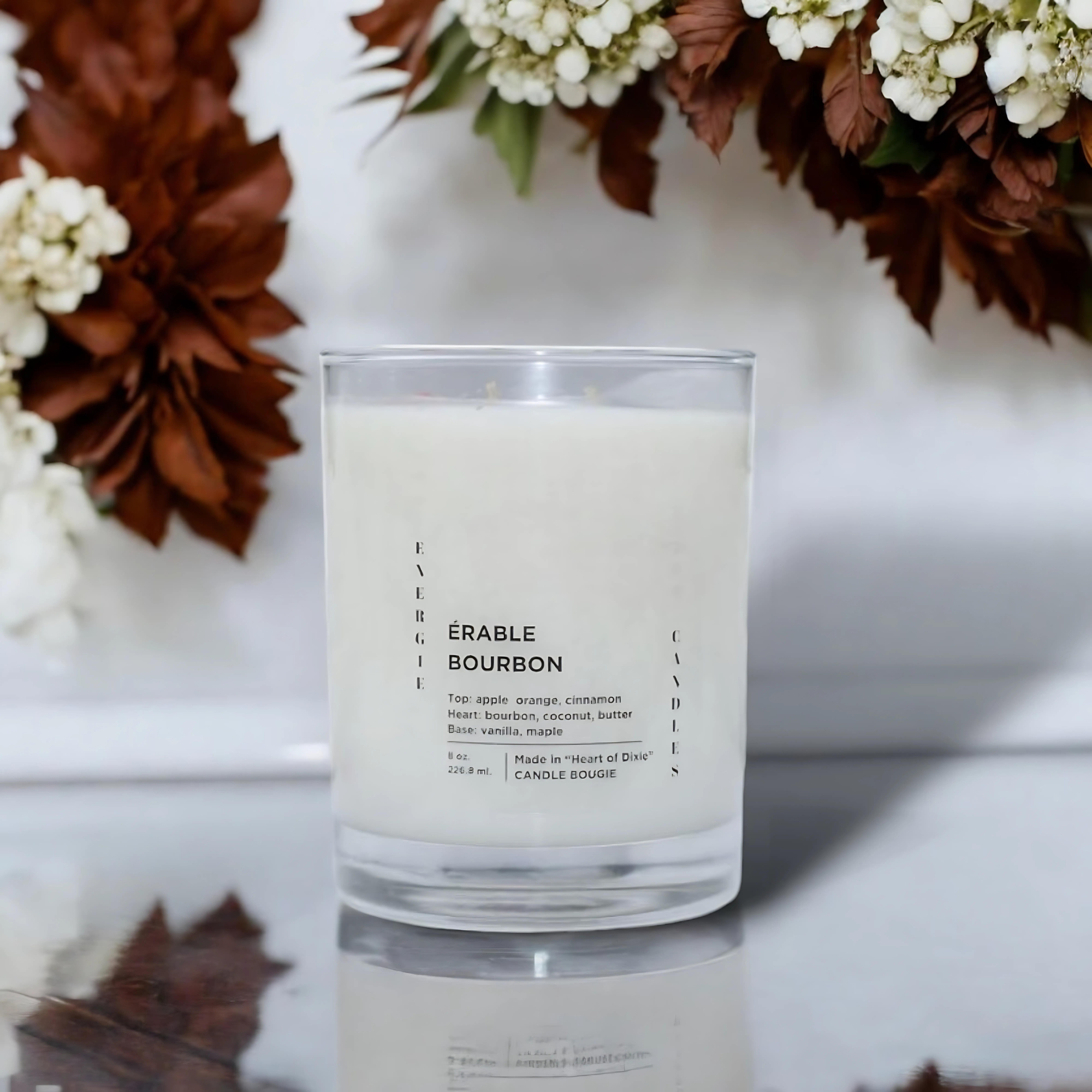 White candle with a cotton wick on a white table, surrounded by brown leaves and white ivy plants, with a white background.