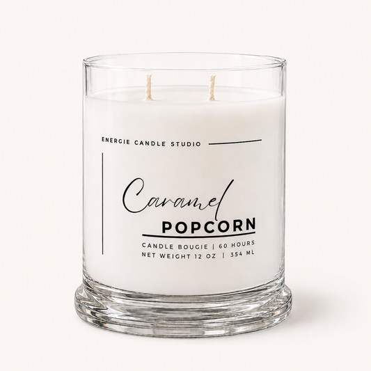 A two-wick candle in a clear glass jar with a thick base, labeled 'Caramel Popcorn' by Energie Candle Studio, on a light cream background.