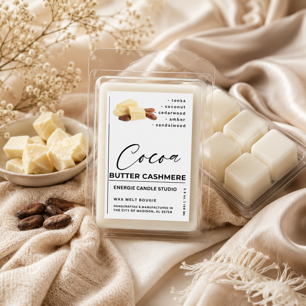 A packaged wax melt candle product labeled 'Cocoa Butter Cashmere' by Energie Candle Studio, shown with a clear clamshell package and a second clear tray of white wax cubes, styled on beige satin fabric with dried flowers and wax chunks nearby.