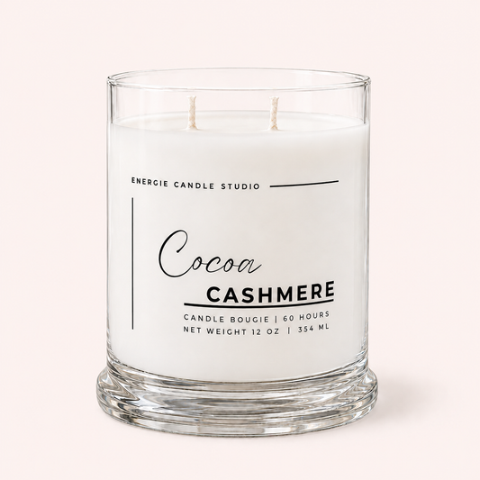 A two-wick candle in a clear glass jar with white wax, labeled 'Cocoa Cashmere' by Energie Candle Studio, set against a solid pale pink background.