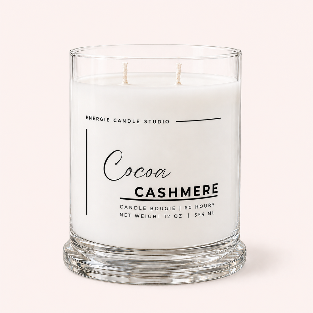 A two-wick candle in a clear glass jar with white wax, labeled 'Cocoa Cashmere' by Energie Candle Studio, set against a solid pale pink background.