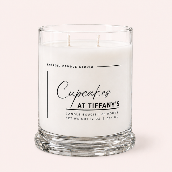 A two-wick candle in a clear glass jar with white wax, featuring a printed label that reads 'Cupcakes AT TIFFANY'S' and 'ENERGIE CANDLE STUDIO'. The candle is photographed against a solid, pale pink background.