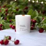 A white candle with a wooden wick sits amidst ripe, tart cranberries and green leaves, with a soft, natural background that highlights the freshness and warmth of the seasonal scent.