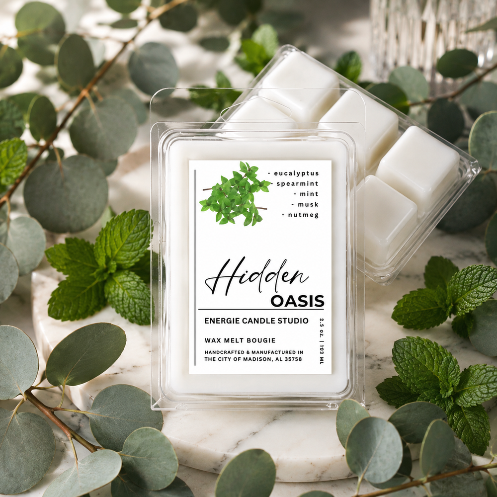 A clear plastic clamshell package of Hidden Oasis wax melt bougie sits on a white marble surface, surrounded by eucalyptus leaves and mint leaves. A second package is partially visible behind it, showing white wax cubes inside.