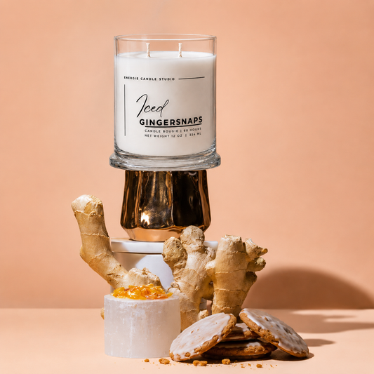 A glass jar candle labeled 'Iced Gingersnaps' by Amber Candle Studio, styled with ginger roots, cookie crumbs, and iced cookies on a peach background.