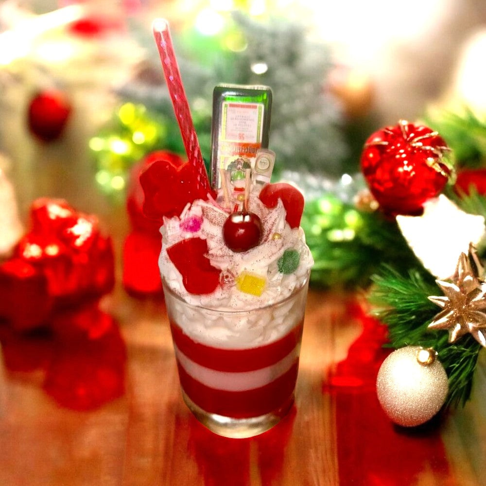 Red and white striped candle encased in a glass vessel with white wax and whipped cream topped with a wax cherry, candy cane, gum drops, and a straw, alongside a mini bottle of Jägermeister. The candle is placed on a brown table with green and red Christmas decorations.