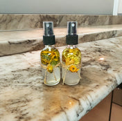 Two clear bottles with black tops filled with yellow botanicals, placed on a two-tier brown marble shelf with a gray wall in the background.