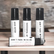 Four bottles of roll-on black oils with white labels, arranged on a wooden table with a blurred gray background.