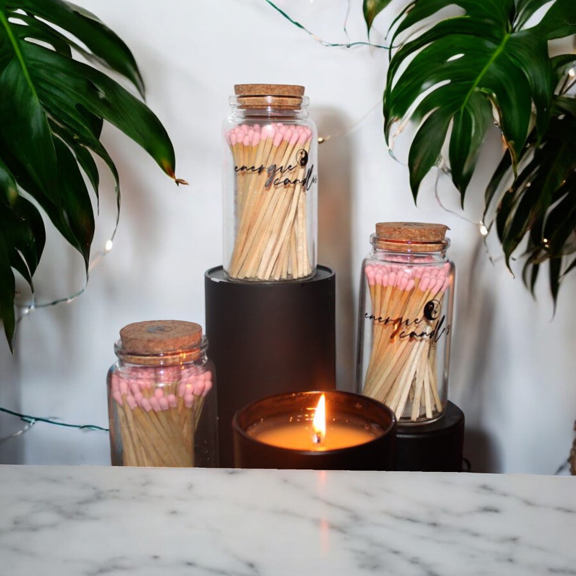 Three glass jars with cork tops filled with wooden matches with baby pink tips, sitting on a black platform with a candle in front. The scene is surrounded by green plants and string lights, placed on a marble table with a white background