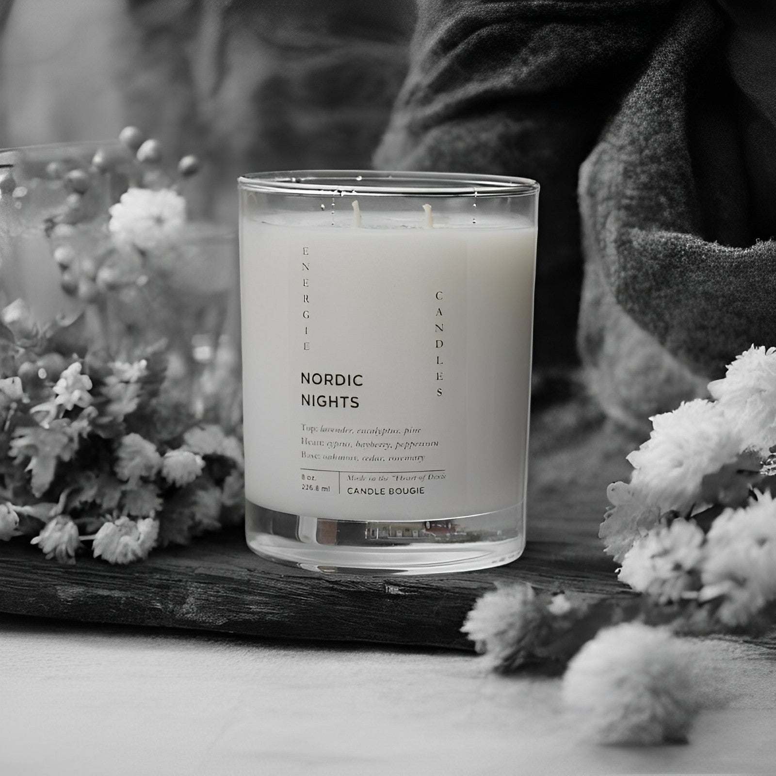 Glass candle with two wicks sitting on a brown stone, surrounded by multiple white flowers, with a brown towel in the background. The entire image is in black and white