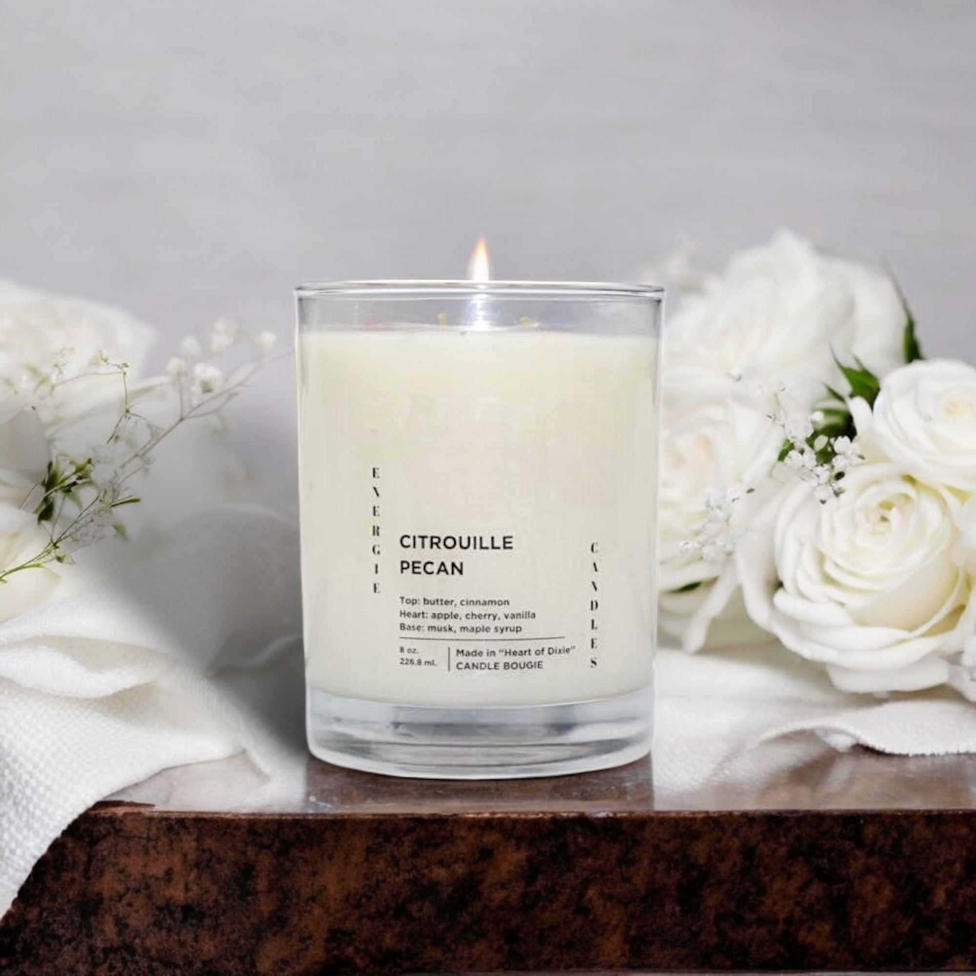 White candle with a cotton wick, encased in a clear glass vessel, sitting on a dark brown marble table. Large white flowers and a white silk cloth create an elegant background.