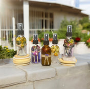 Four clear glass bottles and one amber bottle filled with botanical flowers, arranged against a white brick wall with a lush green garden and house in the background.