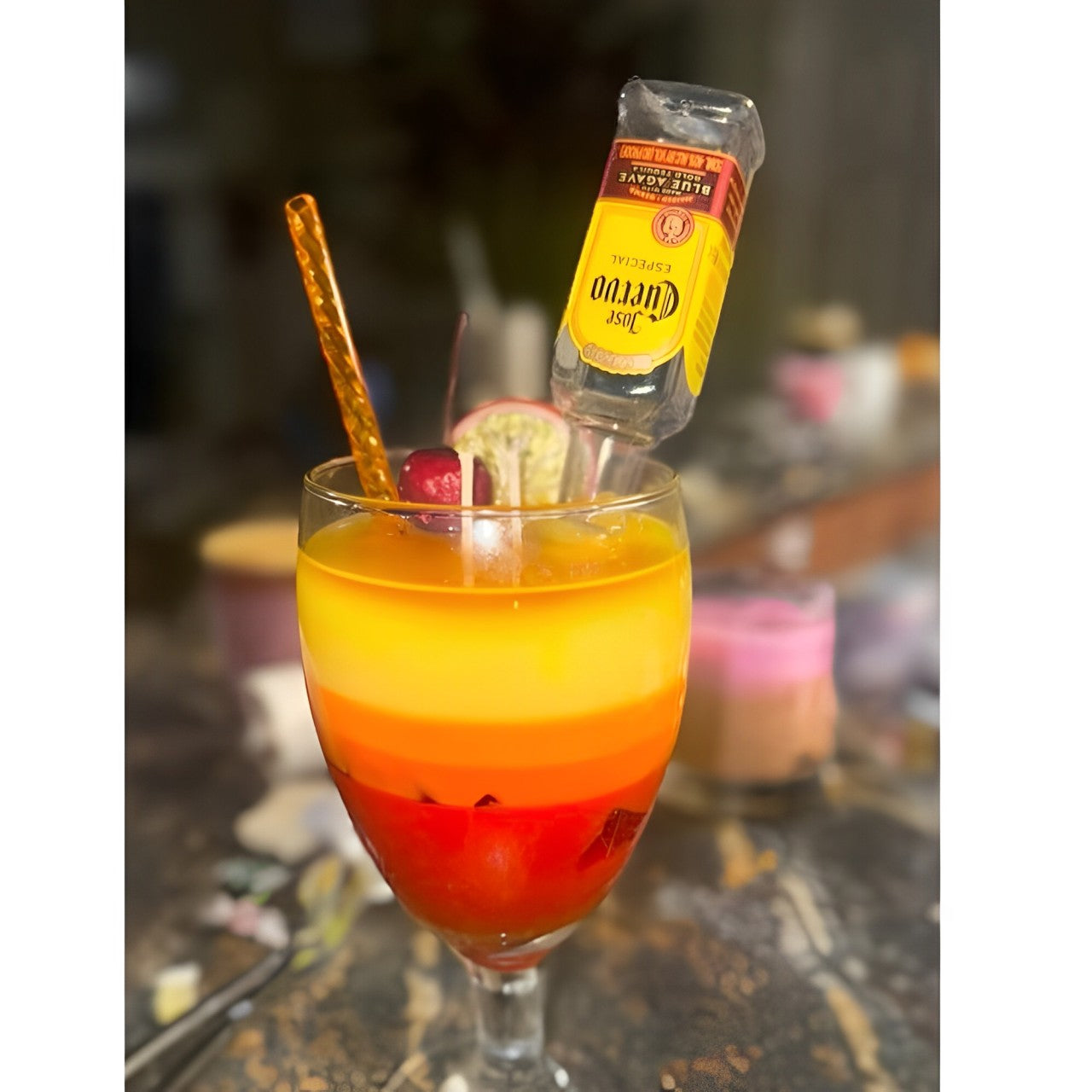 Dessert glass with red, orange, and yellow candles, topped with a red cherry and orange straw, next to a mini bottle of Jose Cuervo tequila on a brown marble table with a blurred background