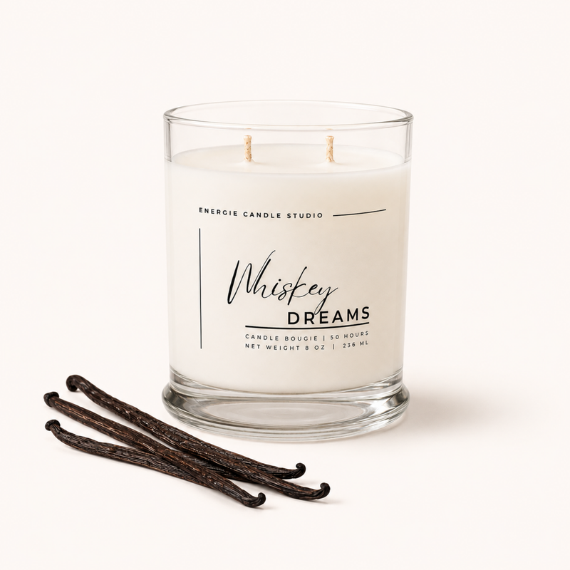 A two-wick scented candle in a clear glass jar with white wax, labeled 'Whiskey Dreams' by Energie Candle Studio, shown on a light beige background with three vanilla beans in front.