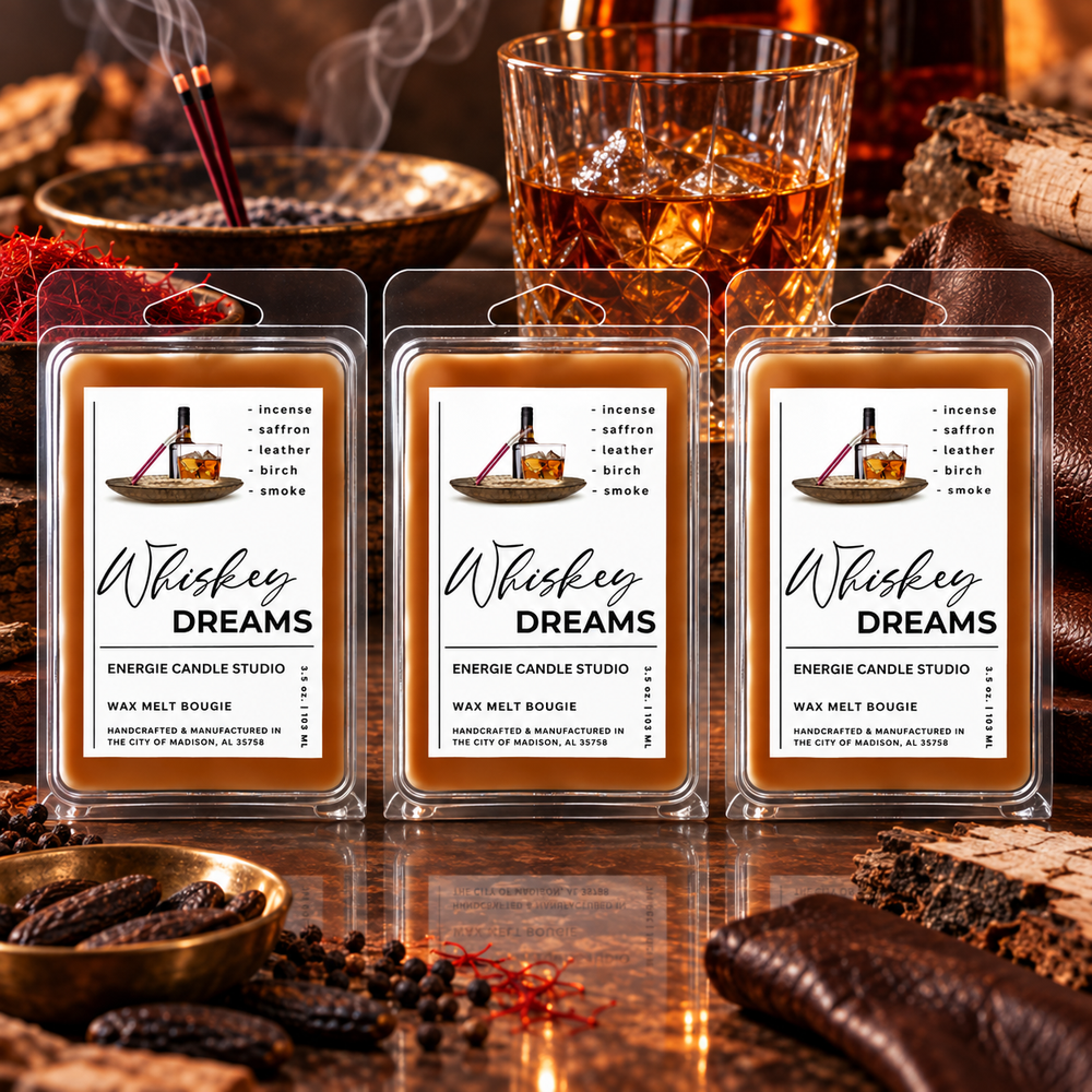 Three packaged wax melts labeled 'Whiskey Dreams' by Emergie Candle Studio are arranged on a reflective surface with whiskey glassware, dried herbs, and wood in the background, creating a warm, smoky bar-like setting.