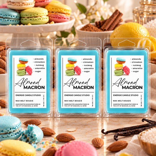 Three packaged wax melts labeled Almond Macron are arranged on a tabletop with macarons, almonds, cinnamon sticks, and vanilla beans around them. The product packaging has a bright blue label with macaron imagery and the brand name Energie Candle Studio.