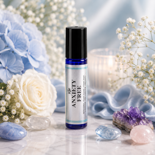 A blue glass roller bottle labeled 'ANXIETY FREE' stands on a marble surface surrounded by white and blue flowers, crystals, and soft blue fabric in a styled product scene.