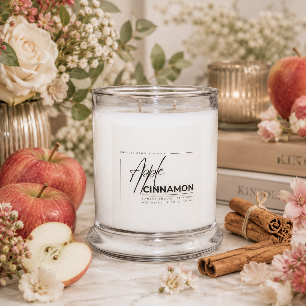 A white soy candle in a clear glass jar labeled 'Apple Cinnamon' by Sweet Scents Studio, styled on a marble surface with red apples, cinnamon sticks, flowers, and books in the background.
