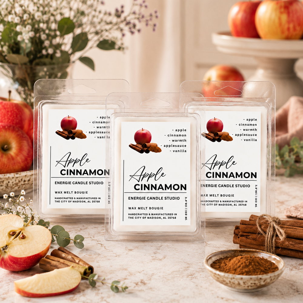Three clear plastic wax melt packages labeled Apple Cinnamon are arranged on a light surface with sliced apples, cinnamon sticks, and a small bowl of cinnamon powder around them. The packages show the brand Energie Candle Studio and include a red apple and brown cinnamon illustration on the labels.
