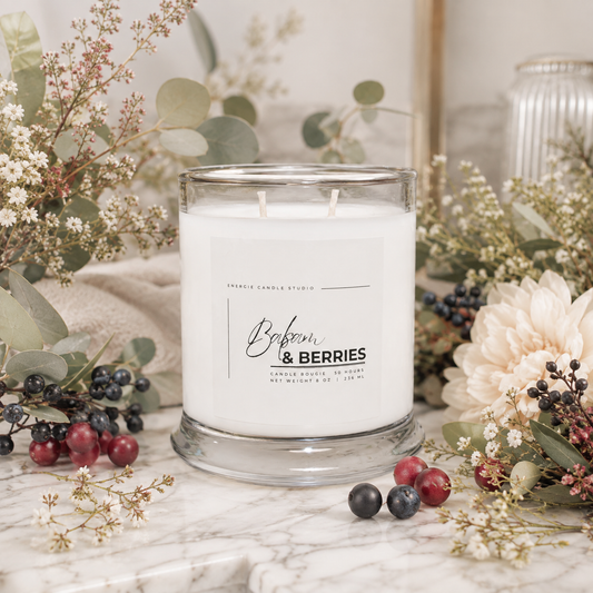 A white scented candle in a clear glass jar sits on a marble surface surrounded by dried flowers, greenery, and berries. The front label reads 'Babyn & BERRIES' with 'Candle Studio' above it.
