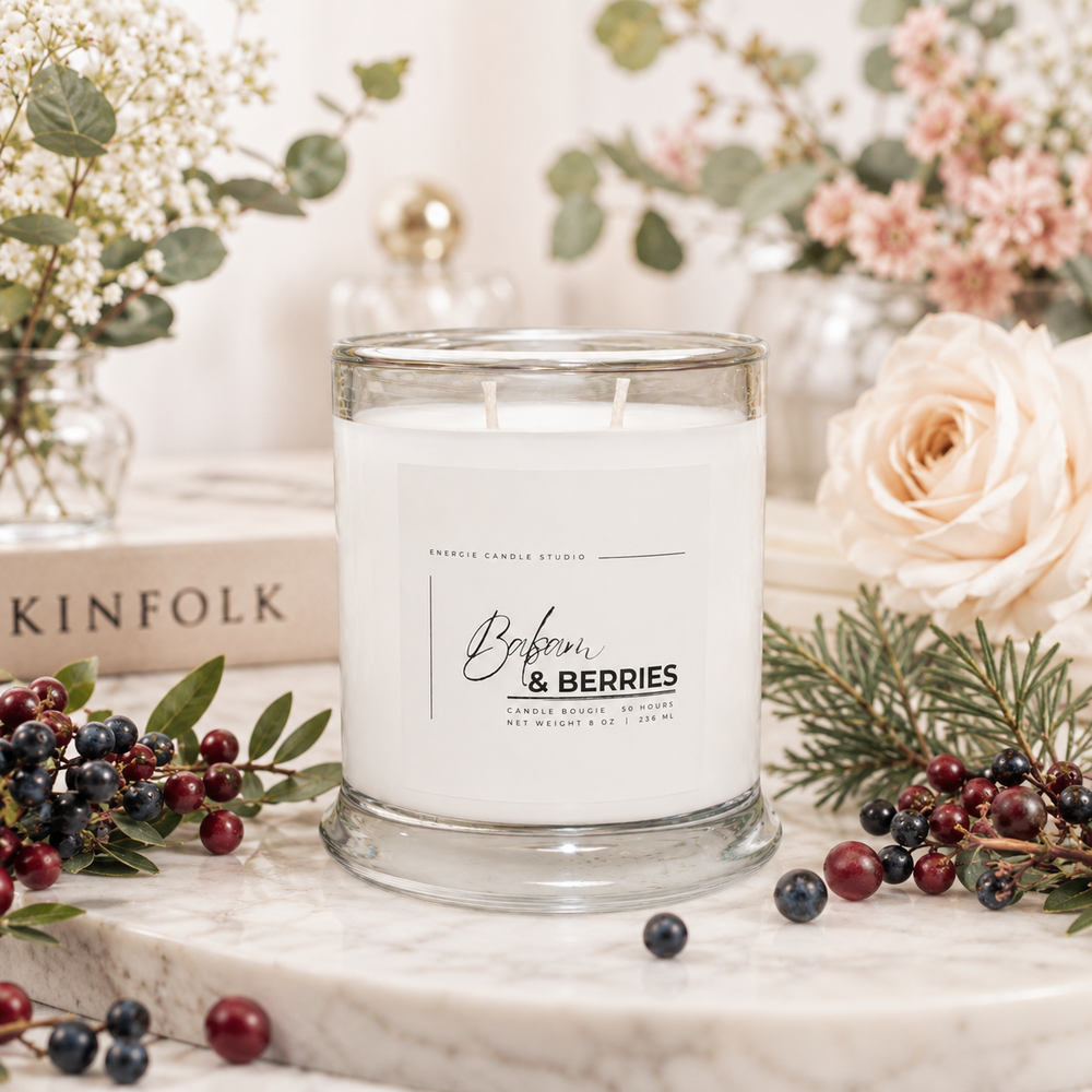 A white candle in a clear glass jar with a white label reading 'Bibberries & Berries' by Sacred Candle Studio, styled on a marble surface with berries, greenery, flowers, and a book in the background.