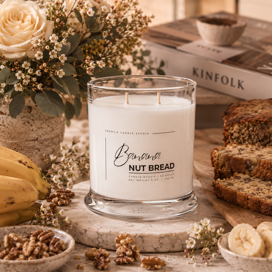 A glass jar candle labeled 'Banana Nut Bread' by Candlestick Candle Studio, styled on a marble board with bananas, sliced bread, walnuts, and dried flowers in a warm, rustic setting.