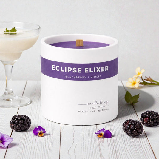 Candle labeled 'Eclipse Elixer' with blackberries and flowers on a wooden surface